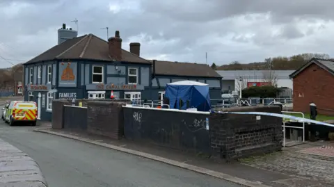 A blue tent on a canal bridge, with police tape stretching away to the right. A police forensics van is parked nearby alongside a pub