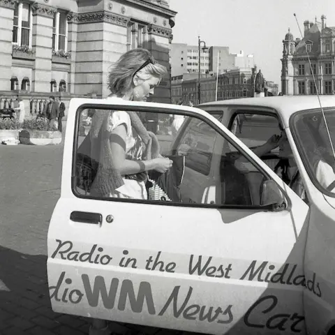 Alan Van Wijgerden A blonde woman stands behind the door of a vehicle branded Radio WM, Radio in the West Midlands