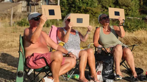 Getty Images Three people sitting on camping chairs hold what looks like pieces of black glass in frames of cardboard to the sky to watch the partial eclipse
