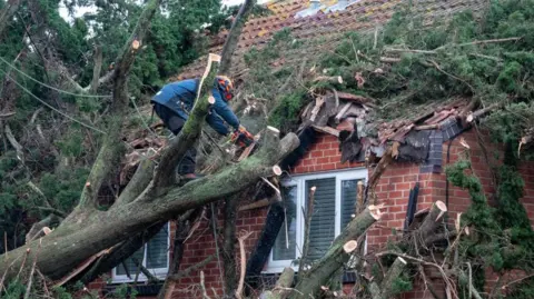 Getty A contractor wearing a hard hat uses a chainsaw to clear a tree that has fallen on the tiled roof of a house in Burnham-on-Sea after high winds in December 2024. 