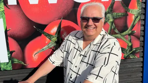 Tony Johnson is smiling at the camera and wearing a white shirt with black petterns on it.