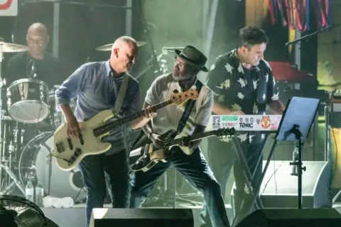 Getty Images Bassist Horace Panter can be seen playing a white-bodied bass, with guitarist Lynval Golding seen on stage next to him. Terry Hall can also be seen on stage playing keyboards. 