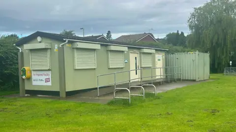Dean Mitchell A long green shed with a green shipping-style container at the far end. It is sitting on concrete at the egde of a grassy area.