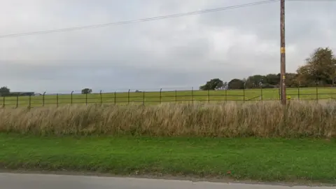 Google The picture shows a quiet, open stretch of countryside beside a road. In the foreground there is a strip of short green grass next to the edge of the tarmac, followed by taller, rough grass and weeds growing along the roadside verge. Behind that runs a metal fence made up of vertical posts and horizontal rails, separating the road from a wide grassy field.