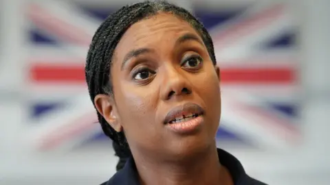 Kemi Badenoch speaking, with a blurred Union Jack in the background