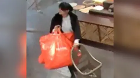 CPS Kaur wearing a grey top and black jacket, captured on CCTV walking through a shop pushing a shopping cart and carrying two large orange bags