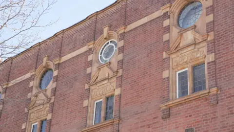 City of Wolverhampton Council The upper section of Wolverhampton Central Library's red brick façade, photographed from below against a clear blue sky. Three first-floor windows with leaded glass are framed by ornate yellow terracotta surrounds, each bearing a poet's name - Shelley, Byron and Spenser. 