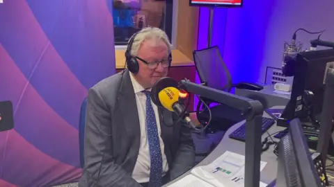 A man with grey hair, glasses and a grey suit, sat in a purple radio studio