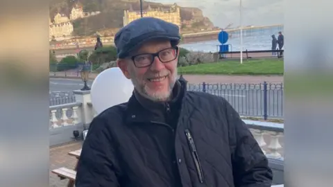 Mr Burke is standing in a cap, glasses and black coat with a coastal scene and pier behind him.