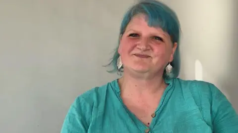 Woman with blue hair and blue top looking into camera. She has feather silver earrings in. 