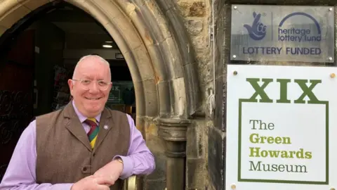An older man, wearing a pink shirt, purple, yellow and green striped tie, brown tweed gilet and glasses stands outside a stone archway next to a sign for The Green Howards Museum. Above the museum sign is plaque with that reads: National Lottery Funded.