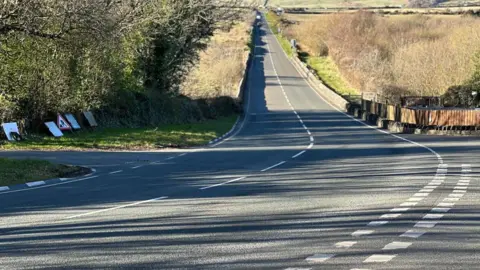 Hillberry Corner, a well-maintained road which has a sharp corner, the road is surrounded by fields.
