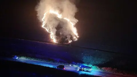 An aerial night-time image showing a concentrated area of burning grass producing flames and a large plume of smoke in a field near Stone Cross. A fire engine with blue lights is parked on a nearby road alongside other vehicles.