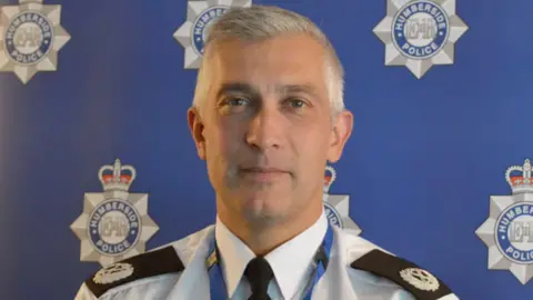 Humberside Police A head and shoulders image of Paul Anderson, the former Chief Constable of Humberside Police. He has grey hair and is wearing uniform consisting of a black tie, white shirt and police lapel badges. He is standing in front of a blue wall featuring multiple Humberside Police logos.