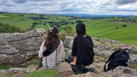 Two young women sat down facing a green field 