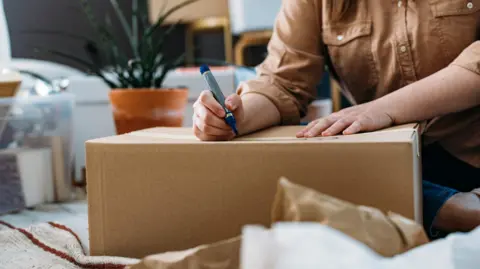 A stock image showing a cardboard box on the floor and a person's hands on top, they are writing with a marker pen on the top of the box. In the background we can see other boxes, packaging and a pot plant.