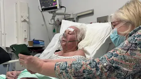 An elderly lady, Mary Lamerton is lying on a trolly in A&E. There is a lady sat next to her in a mask who is holding a phone. 