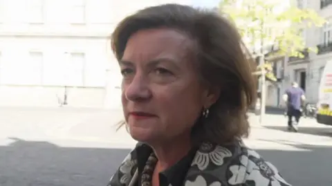 Eluned Morgan arriving at Lancaster House, wearing a grey and white top with flower patterns and a necklace. A man is out of focus in the background, walking away from the camera.