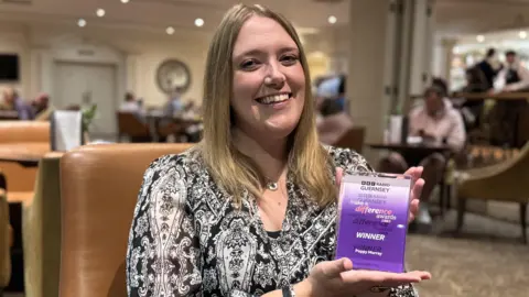 A young woman with shoulder length blonde hair with a middle parting, smiles at the camera. Sitting in a leather chair in a bar, wearing a black and white paisley top, holding a purple and white rectangular award in her hands.