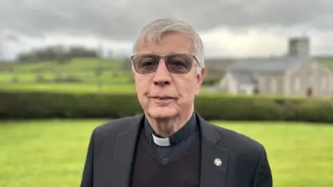 Fr Sean Moore with short grey hair and glasses, he is wearing a black blazer, jumper, shirt and a white clerical collar. In the background is the church with a field and hedge in front of it.