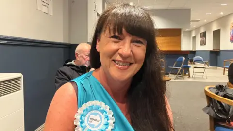 Grace Wood/BBC A smiling woman with long dark hair. She is sat down and is wearing a turquoise top and rosette which reads 'Reform UK'.
