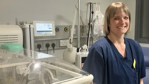 BBC/Katie Radley A woman in a blue hospital uniform smiles at the camera. She is standing next to medical equipment with posters in the background.