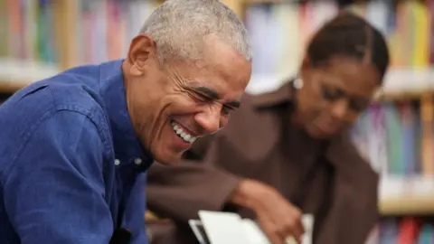 Reuters Image shows Barack Obama, wearing a blue denim shirt, smiling at a child off camera while reading stories to other young people at a library. His wife, Michelle, is out of focus in the background. 