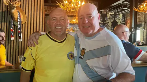 Two people in football shirts standing with arms around each other in a lively bar with antler-style chandeliers, a mounted deer head, and other patrons in the background