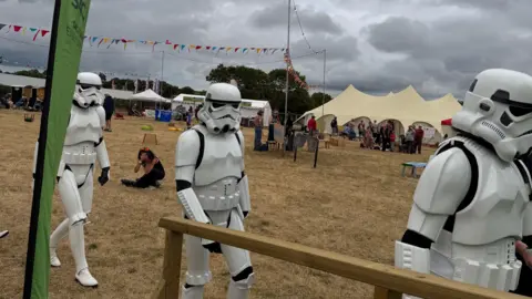 Three Stormtroopers at the National Eisteddfod, walking in a single file line. Tents can be seen in the background on the field where the festival is hosted.