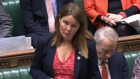 Vikki Slade MP is pictured making a speech in the House of Commons. She has red-brown, straight, long hair and wears a blue jacket and V-necked red and pink top.