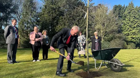 A man with a spade throws earth onto the base of the spine of a tree. A wheelbarrow sits to the side and a number of smartly-dressed figures stand in the background. The sun shines jubilantly on the tall evergreen trees behind them all.