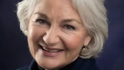 Ellie has short grey hair and is smiling in this close-up photograph. She has earrings in and makeup on and appears with a black background behind her.