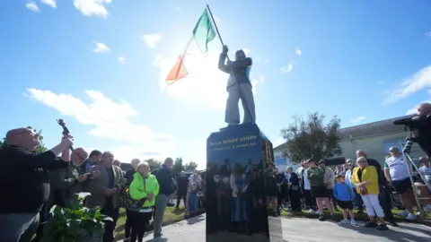 PA Media Members of the public attend the unveiling of the Bobby Sands statue. The statue is in the centre of the photo with crowds of people standing around it. It is a sunny day. 