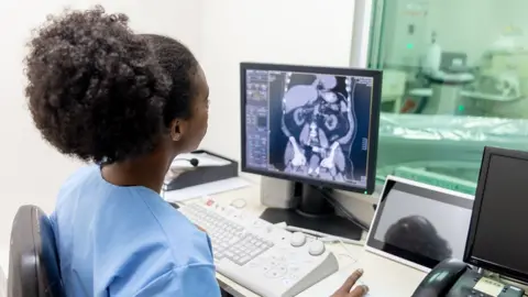 A hospital doctor looking at a medical scan on a computer screen for signs of cancer, with a scan room in the background