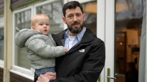 A picture of Thomas DeRonde, standing with his son outside the block of flats where he lives. He is one of the leaseholders affected by an increase in service and sinking fund charges to pay to replace cladding and other fire safety improvements 
