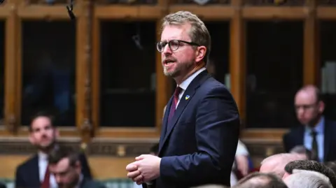House of Commons Joh nSlinger stands and talks in the House of Commons. He wears a dark blue suit jacket and two tone red / purple tie.