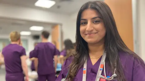 Head and shoulders of Kareena Panesar in purple medical clothing, with other medical students in the background