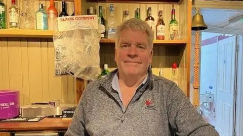 A man standing behind the bar in a pub smiles at the camera. He's wearing a grey fleece. 