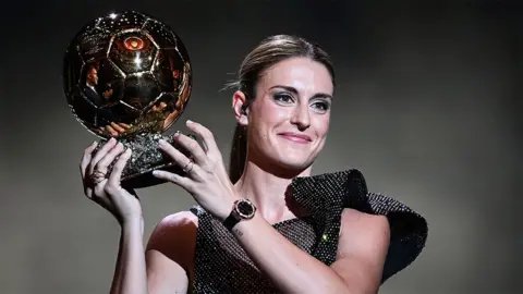 Getty Images Alexia Putellas holds up the soccer ball-shaped trophy, while wearing a glittering black outfit.