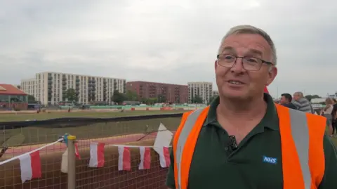 A man with short grey hair and is wearing black glasses. He is wearing a dark green t-shirt and an orange hi-vis jacket. Behind him is a small fence with flag bunting hanging on it. Beyond is a dirt speedway track.