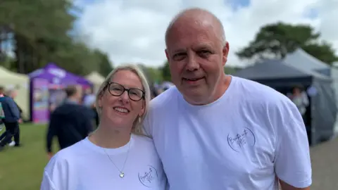 Michael Quine and Julie Kirby wear white T-shirts. He has very short grey hair, she has blonde hair and wears dark-rimmed spectacles.