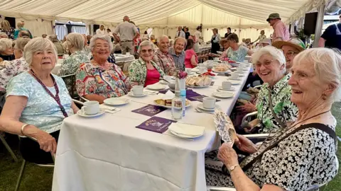 Residents of St Monica Trust villages and care homes gather for an afternoon tea. Several men and women of an older age range sit at a table, with sandwiches and cakes on it. 
