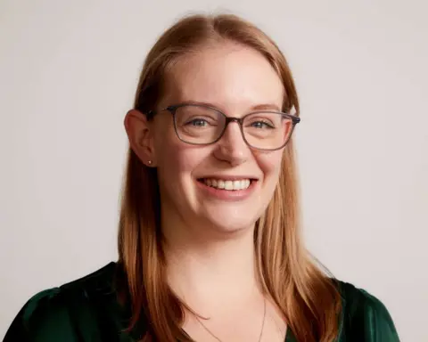 Fran Yarlett A woman with long blonde hair and glasses smiles for a photo in front of a plain white wall