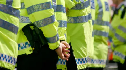The back of a line of police officers, wearing yellow hi-vis uniforms and black trousers. 