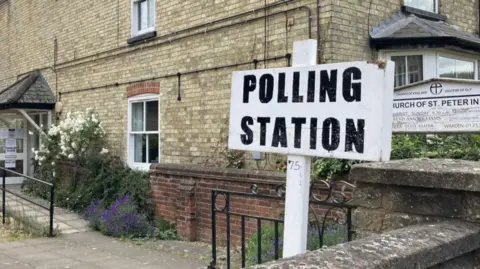 A black writing and white background sign says "polling station" in capital letters, it has been put up outside a church building.