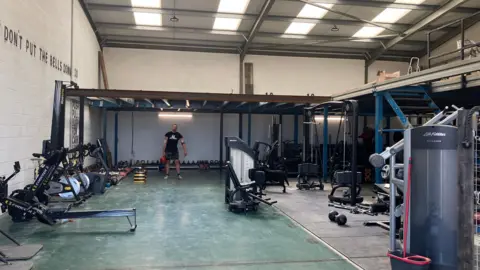 A man is stood under a tall metal frame holding a weight. He is inside a large warehouse type building with exercise equipment around the space