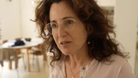 Liat Zvi is pictured from the shoulders up, in a domestic setting with a table in the background. She is a dark-haired woman with gold-rimmed glasses.