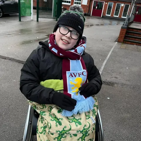 Family A boy smiles at the camera while sitting in his wheelchair. He has a maroon and blue Aston Villa scarf wrapped around his neck and has a yellow blanket with green lizards on it around his lap. He wears a black coat and hat and is sat outside on tarmac, in front of a redbrick building.