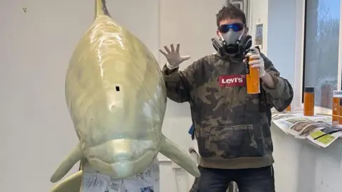 Durlston Country Park A person in a brown camouflage hoodie wearing a respirator facemask posing for the camera next to the dolphin which has its nose facing towards the camera. The person is holding up a spray can and has their other hand held up. The dolphin is covered in pale cream coloured primer.