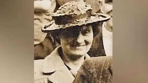 Avon & Somerset Police Unclear, sepia photo of a woman smiling, wearing a brimmed hat and coat